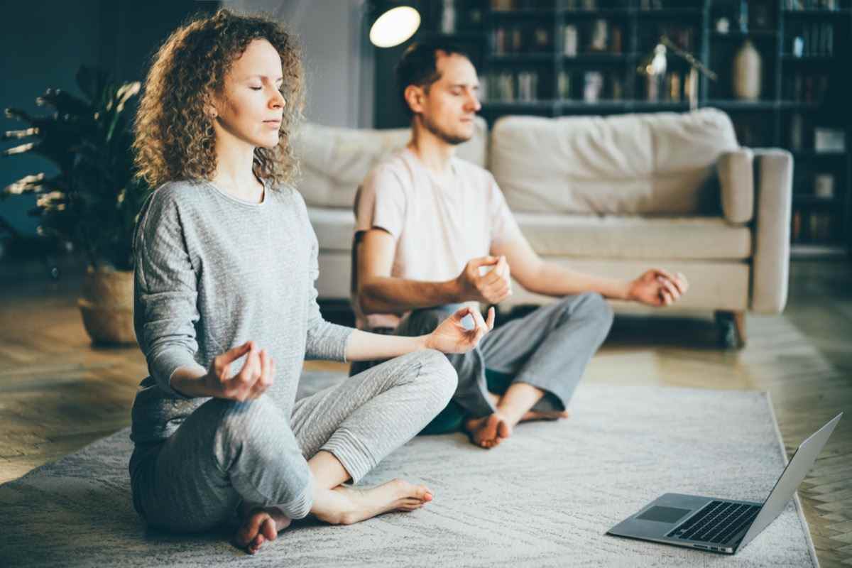 Ruhiges Paar im Pyjama, das meditiert, spirituelle Übungen auf dem Laptop hört, zu Hause auf Lotus-Pose sitzt