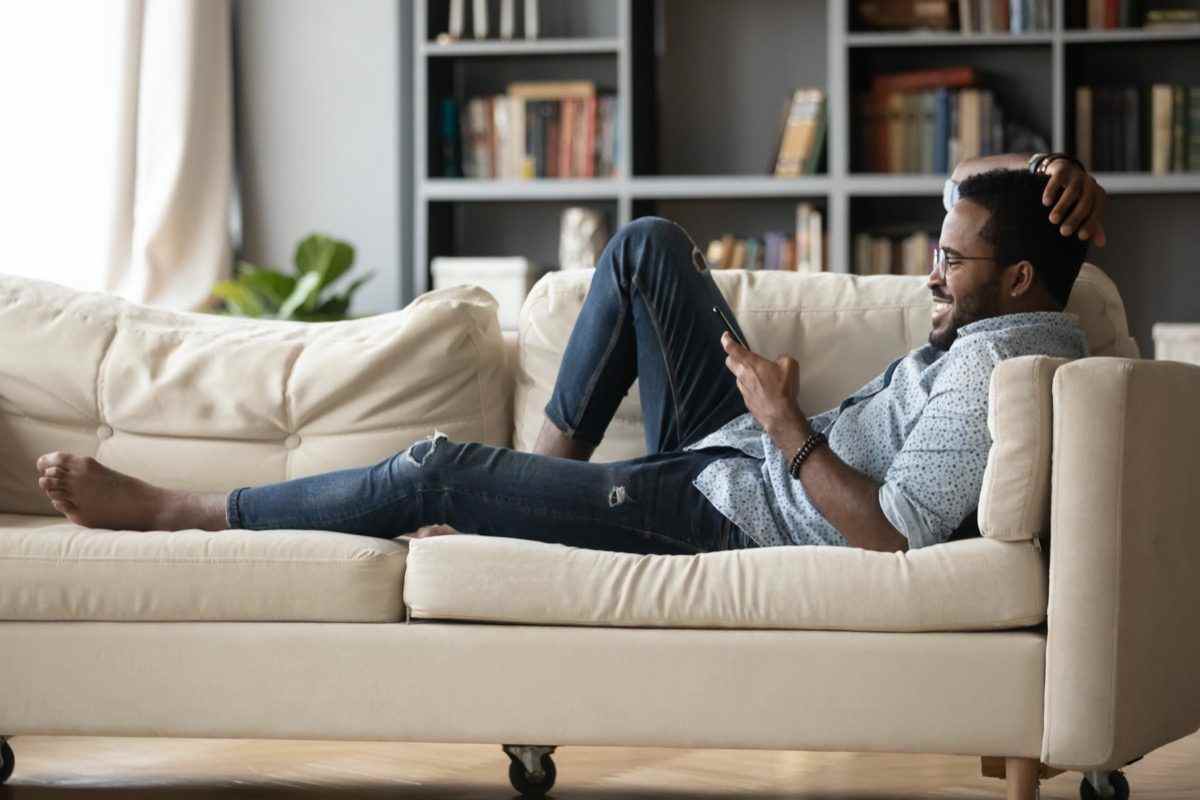 Junger schwarzer Mann liegt auf der Couch und liest Tablet