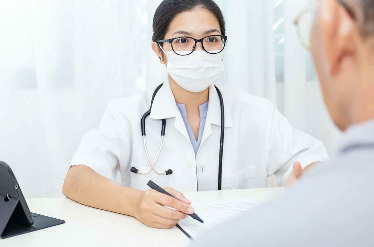Porträt einer asiatischen Ärztin, die eine Schutzmaske trägt, die einem Patienten einige Informationen über die digitale Tablet-Zwischenablage zeigt, der Patient hört dem Facharzt im Klinikbüro zu