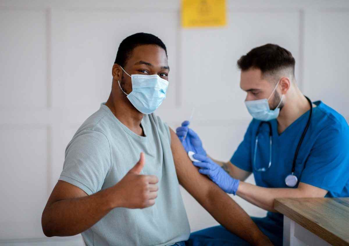 Afroamerikanischer Mann in antiviraler Maske, der während der Coronavirus-Impfung den Daumen nach oben gestikuliert und die Covid-19-Impfung genehmigt