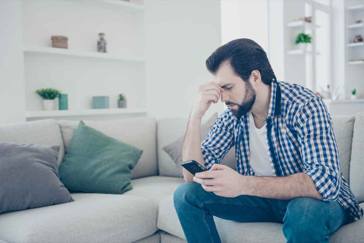 trauriger frustrierter Mann mit Stoppeln in Jeans, Hemd, das Finger zwischen geschlossenen Augen mit Smartphone hält