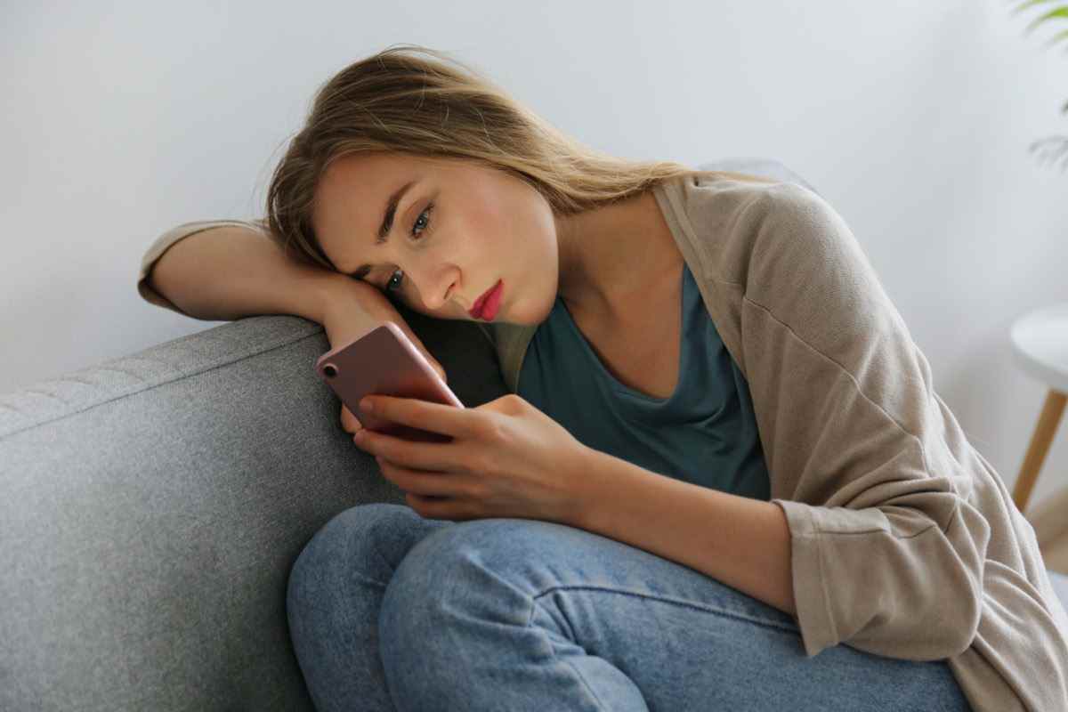Frau mit depressivem Gesichtsausdruck sitzt auf grauer Textilcouch und hält ihr Telefon