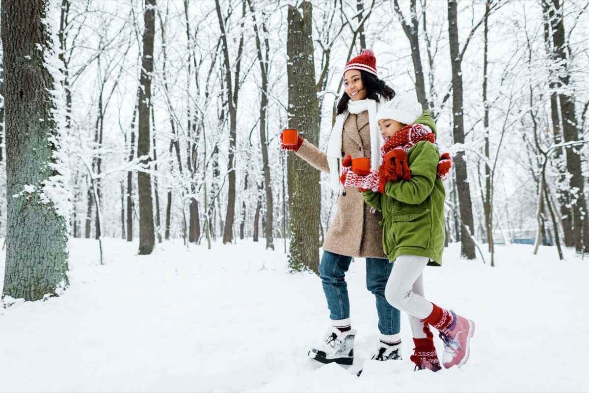Mutter und süße Tochter gehen und halten rote Tassen zwischen Bäumen und betrachten Schnee im verschneiten Wald
