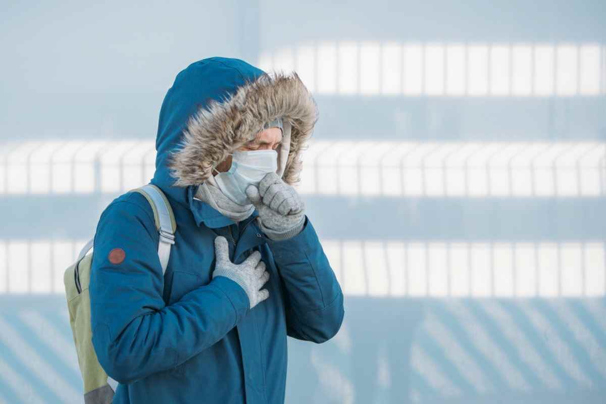 Porträt eines kranken jungen Mannes in blauer Jacke, der eine Kapuze aufsetzt, eine Erkältung hat, sich unwohl fühlt, hustet, eine medizinische Gesichtsmaske trägt, im Freien