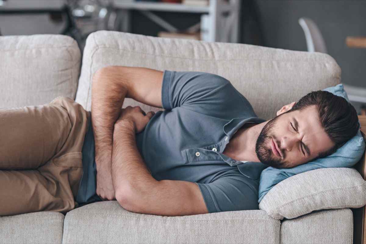 Mann, der seinen Bauch umarmt und die Augen geschlossen hält, während er zu Hause auf der Couch liegt