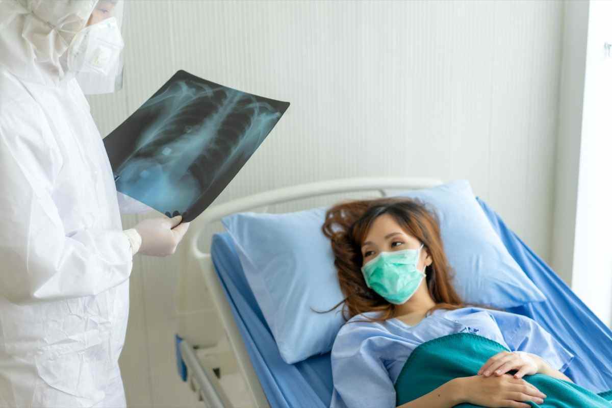 Patienten, die mit Maske auf dem Krankenhausbett liegen, beim Lesen des Ergebnisses des Arztes den Röntgenfilm der Lunge betrachten und eine Behandlung empfehlen