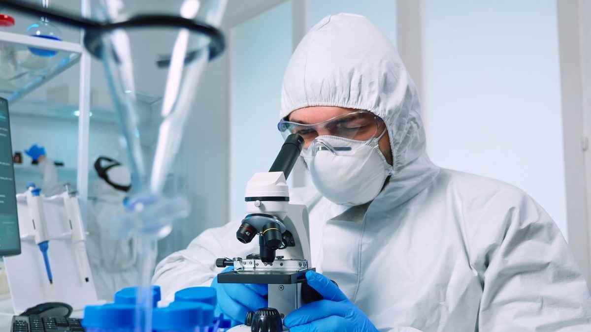 Biotechnologiewissenschaftler im PPE-Anzug, der DNA im Labor unter Verwendung des Mikroskops erforscht.  Team, das die Virusentwicklung mit Hightech für die wissenschaftliche Erforschung der Impfstoffentwicklung gegen Covid19 untersucht