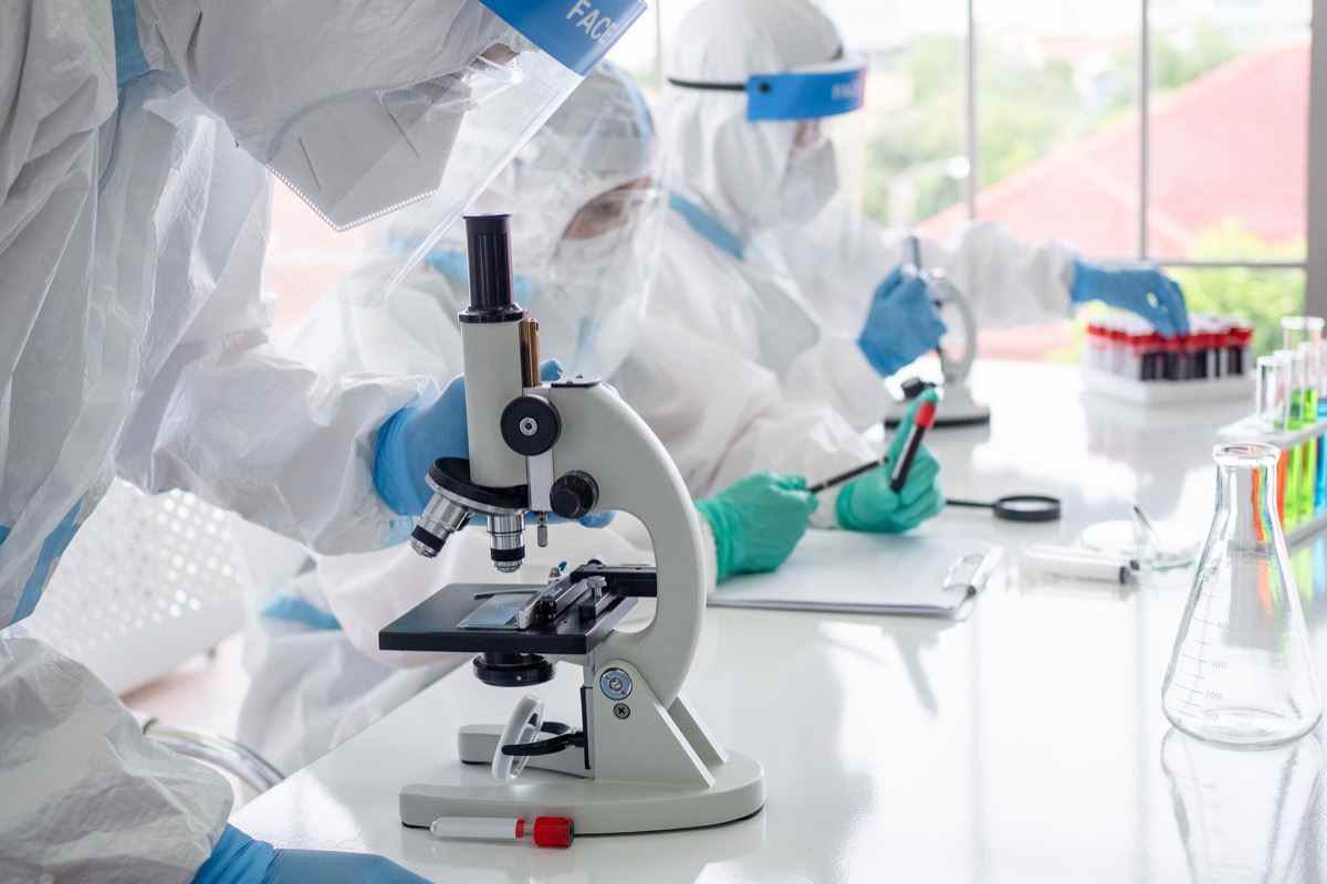 Wissenschaftler und Mikrobiologen mit PSA-Anzug und Gesichtsmaske halten Reagenzglas und Mikroskop im Labor