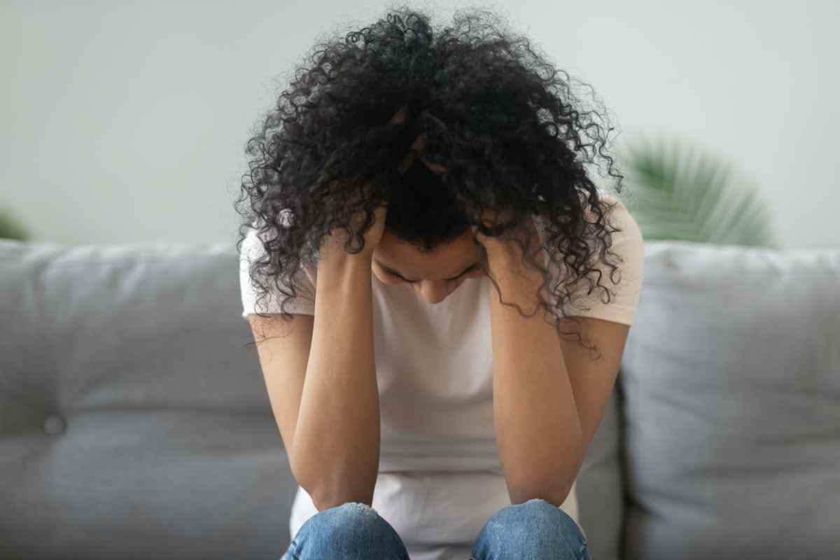 Frau hält den Kopf in den Händen, leidet unter Trauerproblemen, depressives, einsames verärgertes afrikanisches Mädchen, das allein auf dem Sofa zu Hause weint