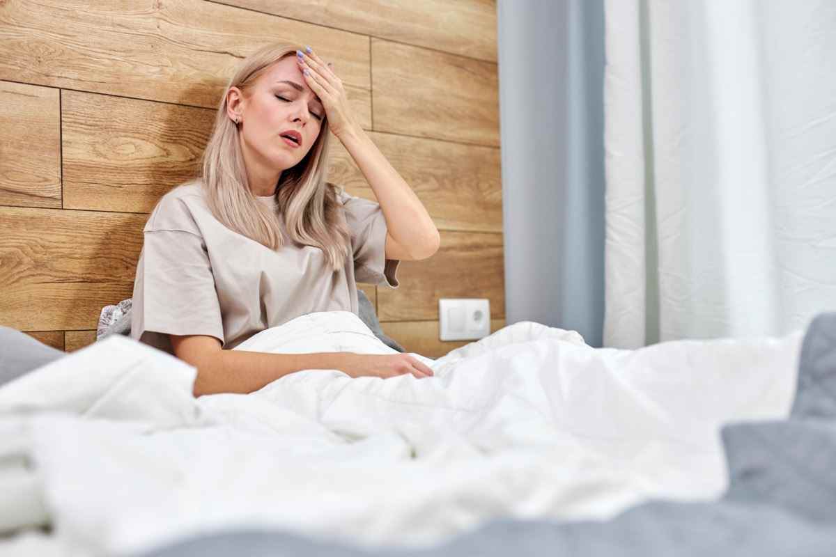 Frau, die krank ist und Grippe hat, die allein zu Hause im Bett sitzt, hohes Fieber oder hohe Temperatur hat und die Stirn berührt