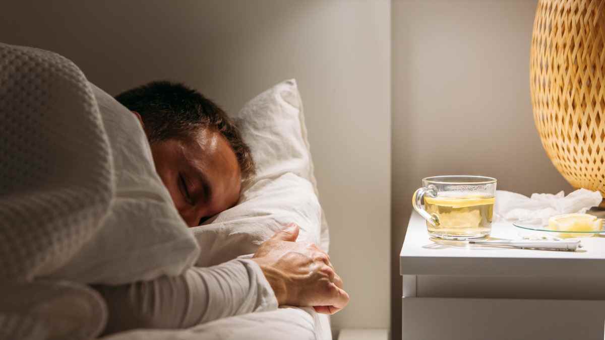 Kranker Mann schläft mit einer Decke bedeckt, die mit hohem Fieber im Bett liegt und sich im Wohnzimmer ausruht.  Er ist erschöpft und leidet an Grippe mit einer Tasse Tee mit Zitrone, Medikamenten.  Grippesaison.