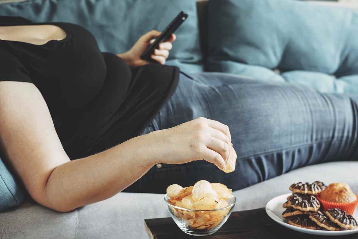 Übergewichtige Frau, die mit dem Smartphone auf dem Sofa liegt und Chips isst