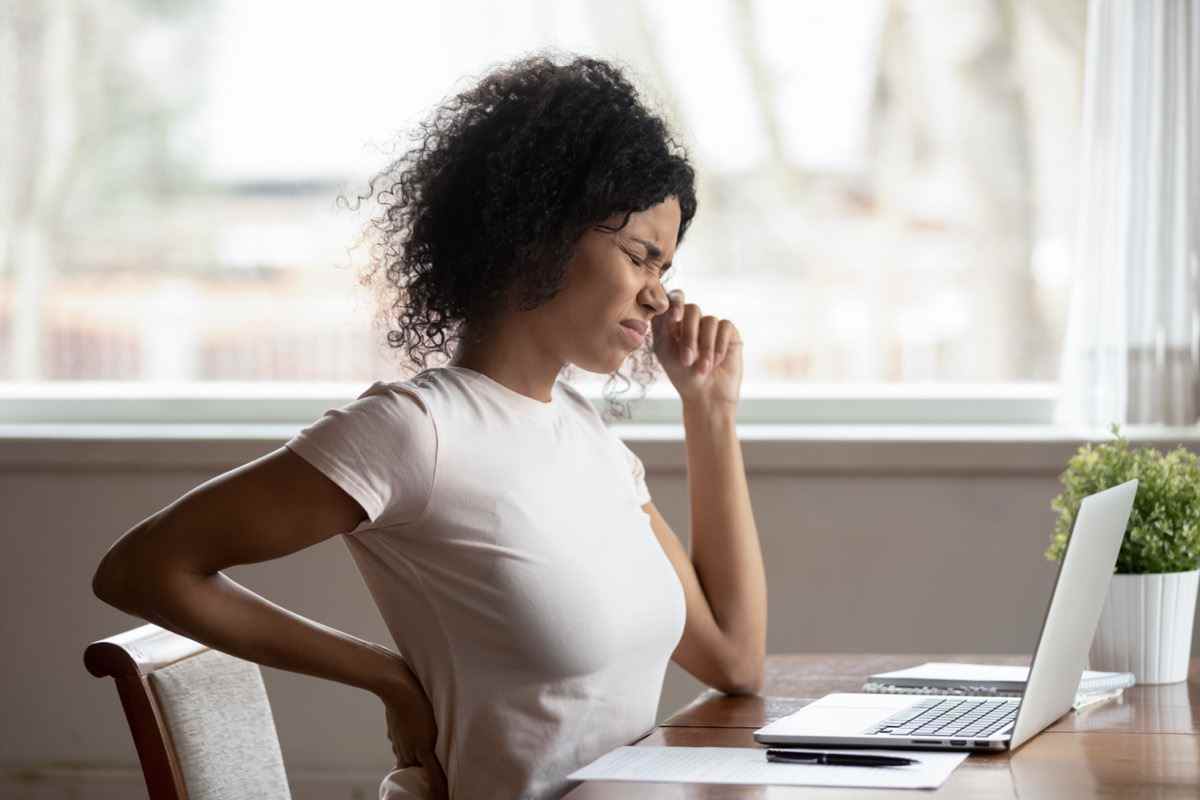 Arbeiterinnen sitzen am Schreibtisch berühren Rücken leiden an unteren Wirbelsäulenkrämpfen, verletzten ungesunde biracial Frauendehnung haben starke Rückenschmerzen.