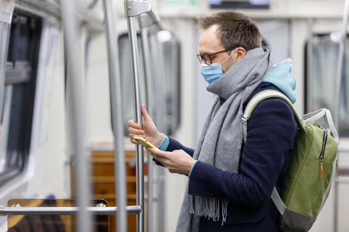 Mann mit Brille, der sich krank fühlt, Schutzmaske gegen übertragbare Infektionskrankheiten trägt und als Schutz vor Grippe in öffentlichen Verkehrsmitteln/U-Bahnen, Smartphone benutzt und betrachtet