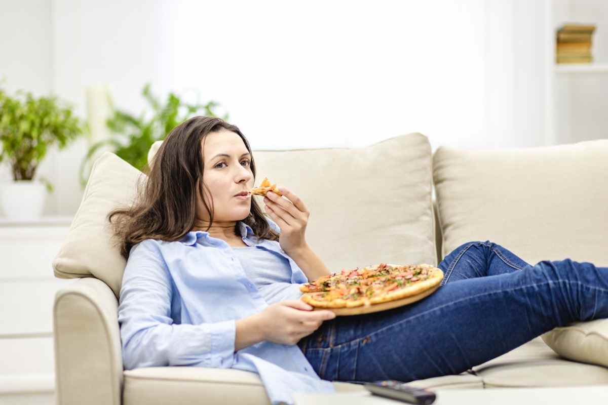 Frau kaut Pizza, während sie auf dem weißen Sofa liegt.  Sie sieht Fernsehsendungen auf unscharfem Hintergrund.