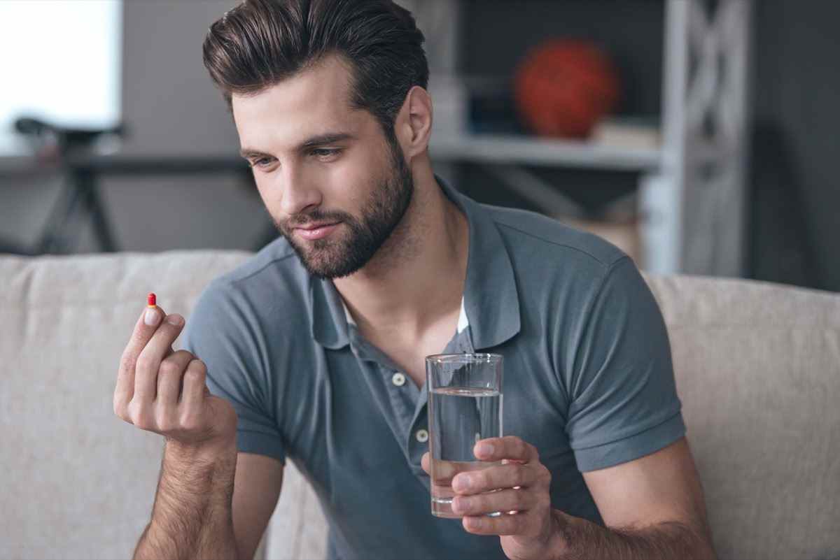 Mann, der ein Glas Wasser hält und eine Pille in der Hand betrachtet, während er auf der Couch sitzt