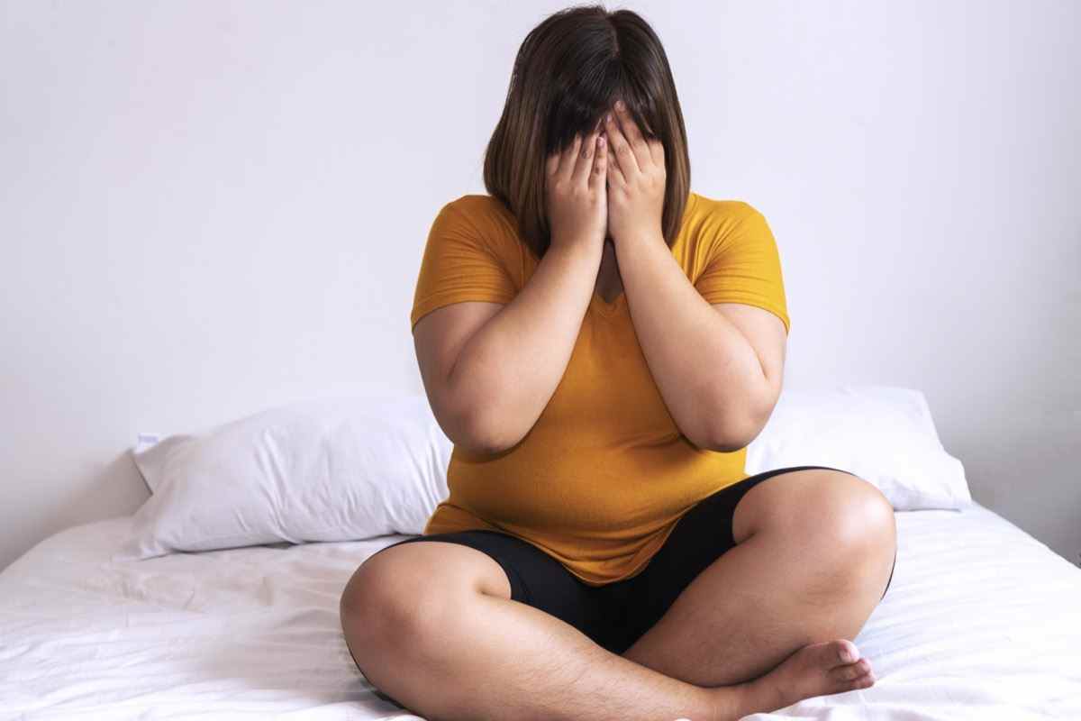 Übergewichtige junge Frau, die auf einem weißen Bett sitzt, während sie zu Hause die Hände auf ihrem Gesicht hält.  Verärgerte Frau, die im Schlafzimmer unter zusätzlichem Gewicht leidet.  Fettleibigkeit ungesundes Konzept.