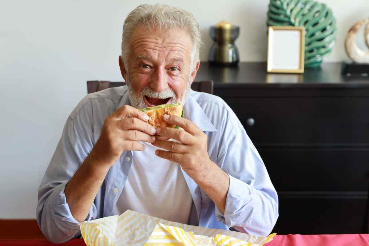 Älterer Mann isst Hamburger im Wohnzimmer mit lächelndem Gesicht