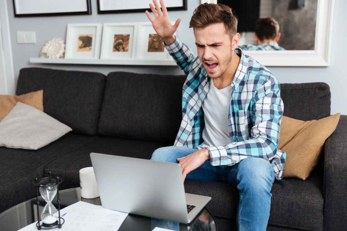 Foto eines wütenden Borstenmannes, der in einem Hemd in einem Käfigdruck gekleidet ist, der zu Hause auf dem Sofa sitzt und einen Laptop benutzt