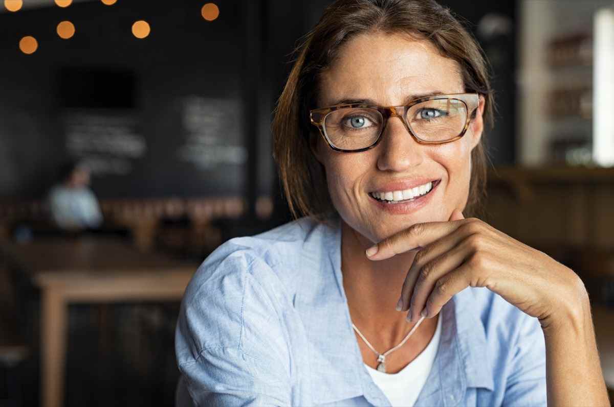 Porträt einer glücklichen reifen Frau mit Brille und Blick in die Kamera.  Nahaufnahmegesicht der lächelnden Frau, die in der Cafeteria mit der Hand am Kinn sitzt.  Erfolgreiche Dame in einem Café-Pub.