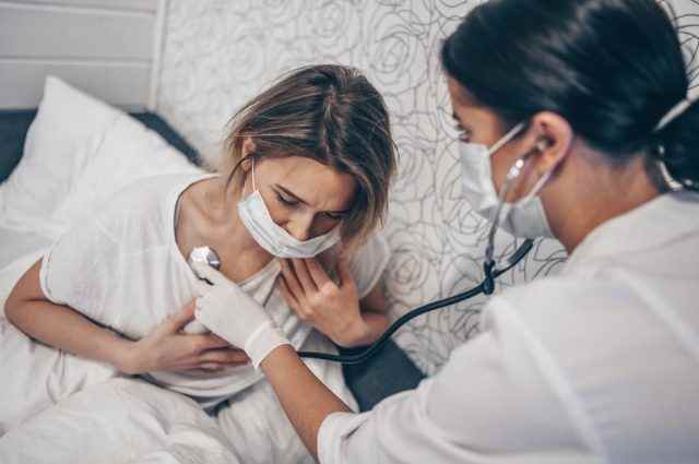 Arztkrankenschwester in schützender Gesichtsmaske, die mit einem Stethoskop atmet, das Coronavirus (COVID-19) vermutet.