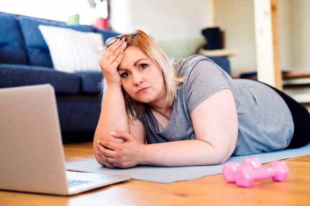 Übergewichtige Frau zu Hause auf dem Boden liegend, Laptop vor sich, bereit zum Training auf Matte laut Video