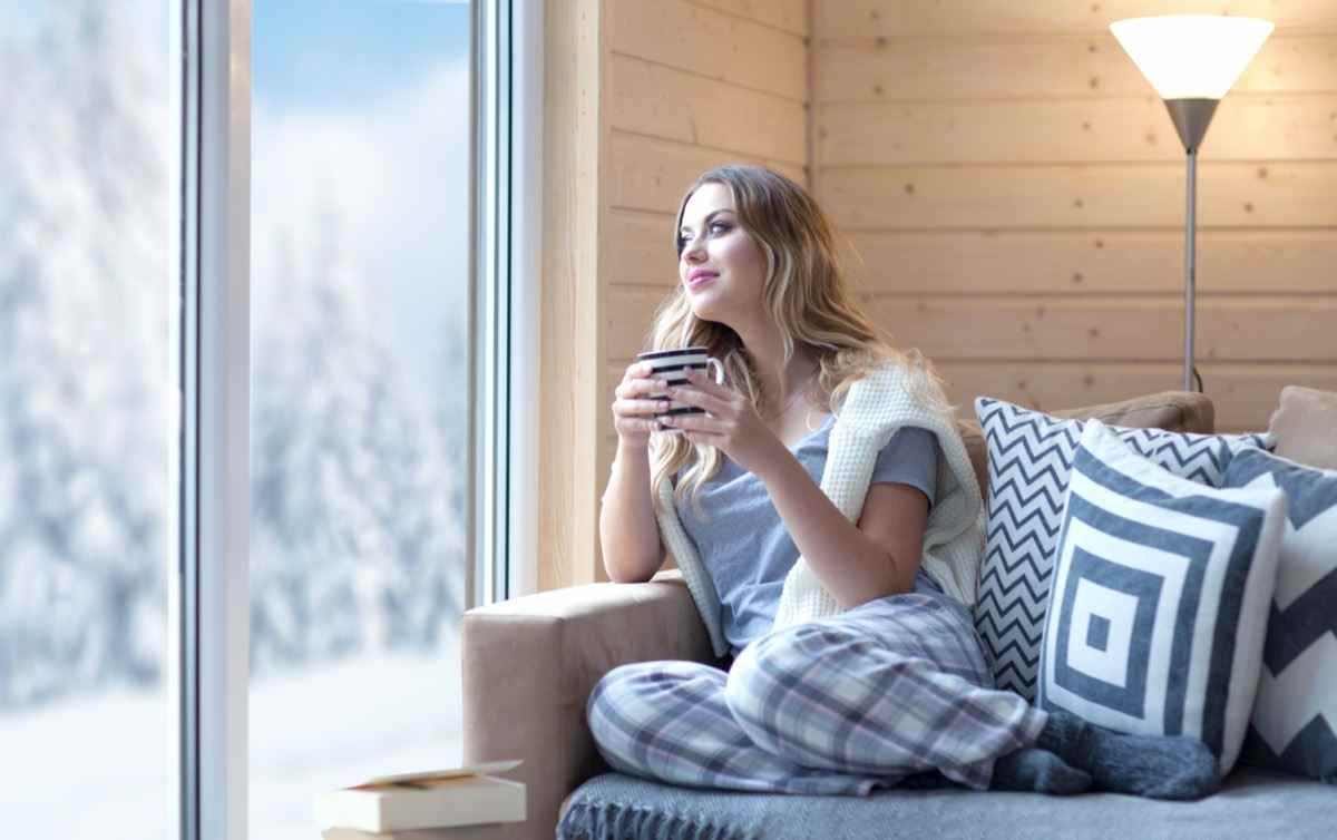 Frau mit Tasse Kaffee zu Hause im Wohnzimmer am Fenster sitzen.  Winterschneelandschaftsansicht