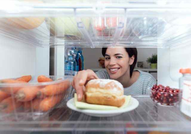 Lächelnde junge Frau, die einen ungesunden Snack isst, nimmt ein köstliches Gebäck aus dem Kühlschrank