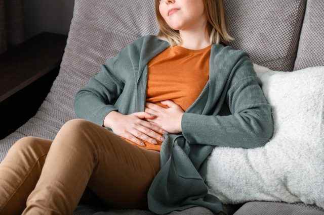 Junge Frau leidet, krümmt sich vor Bauchschmerzen auf der Couch im Wohnzimmer zu Hause liegend