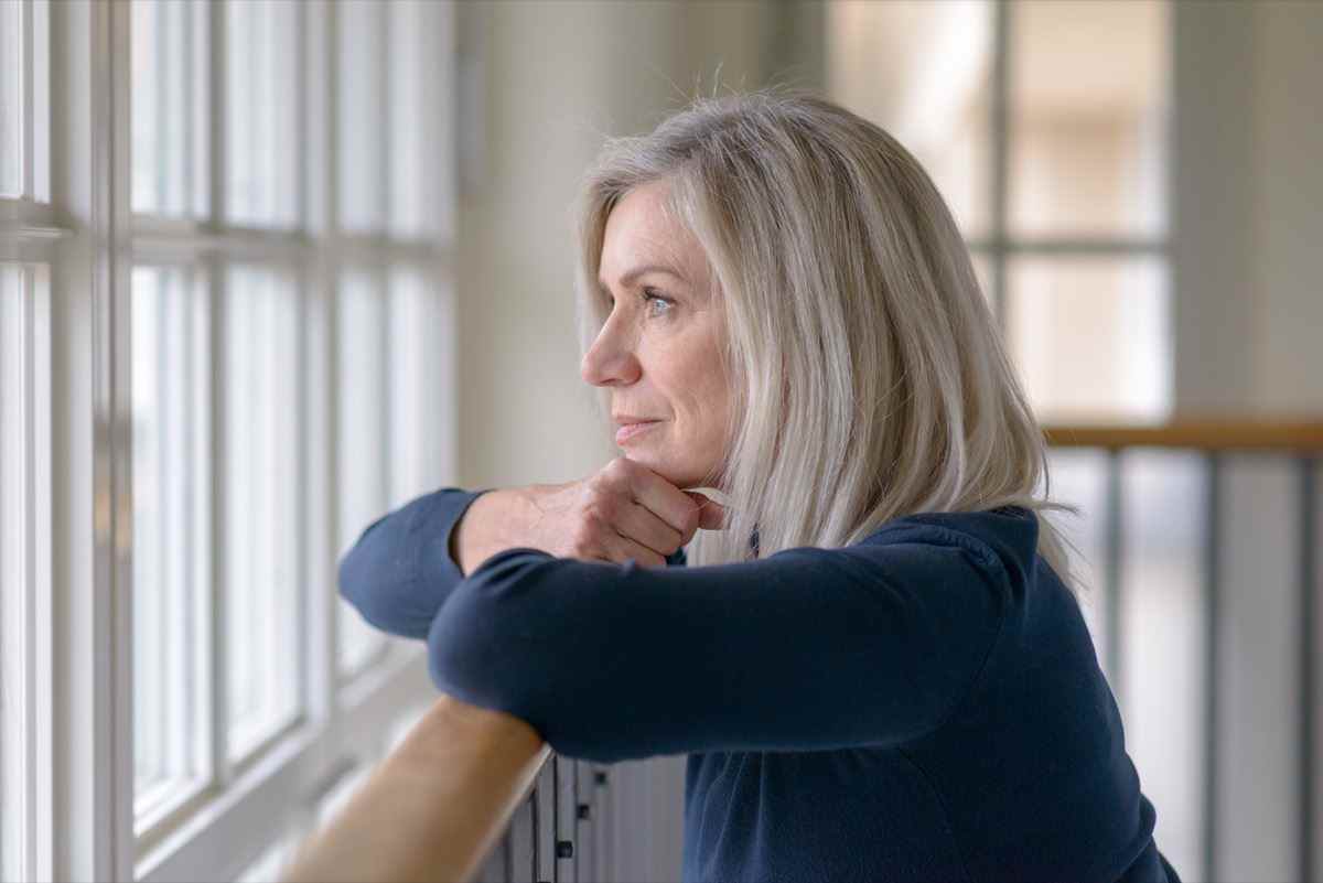 Traurige blonde Frau, die durch ein Fenster mit einem ernsten Gesichtsausdruck beobachtet, der ihr Kinn auf ihre Hände stützt, während sie sich auf ein Holzgeländer stützt
