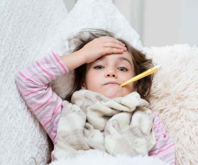 Krankes Mädchen liegt im Bett mit einem Thermometer im Mund und berührt seine Stirn