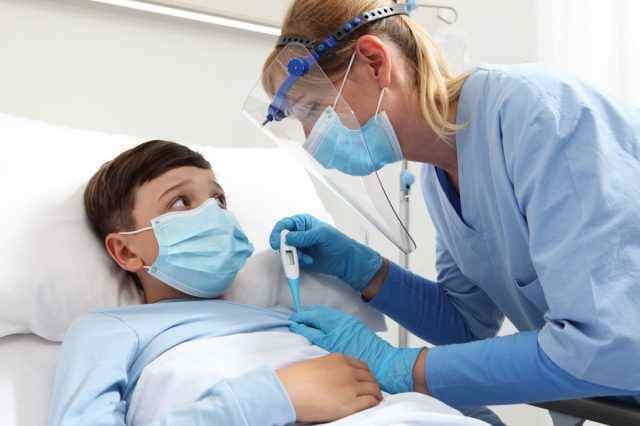 Krankenschwester mit Thermometer misst Fieber beim geduldigen Kind im Krankenhausbett und trägt Schutzvisier und chirurgische Maske.