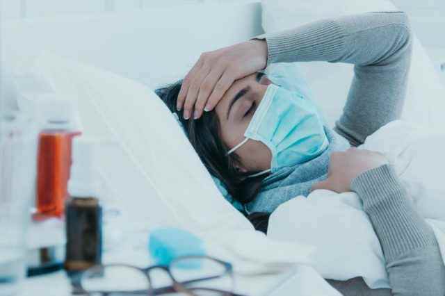 Junge kranke Frau liegt müde mit Gesichtsmaske im Bett und hält sich wegen Kopfschmerzen den Kopf.