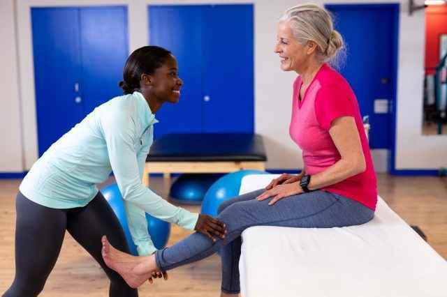 Fröhliche Physiotherapeutin, die einer aktiven Seniorin im Sportzentrum eine Beinmassage gibt