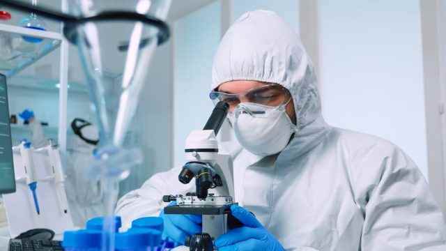 Biotechnologie-Wissenschaftler im PSA-Anzug, der DNA im Labor unter Verwendung des Mikroskops erforscht.  team, das die virusentwicklung mit hightech für die wissenschaftliche forschung der impfstoffentwicklung gegen covid19 untersucht