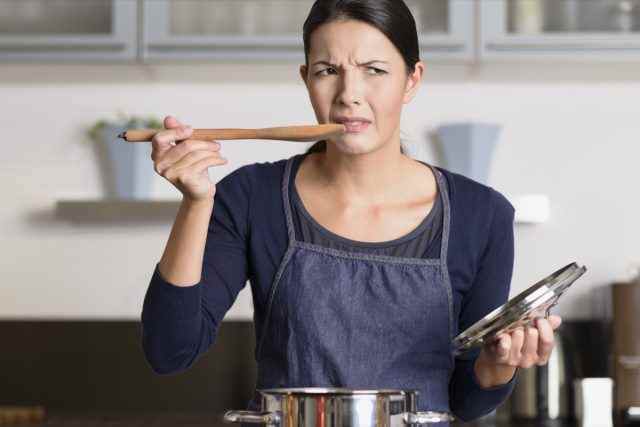 Köchin, die in ihrer Schürze am Kochfeld steht und ihr Essen im Topf mit einer Grimasse probiert, da sie es geschmacklos und ungenießbar findet