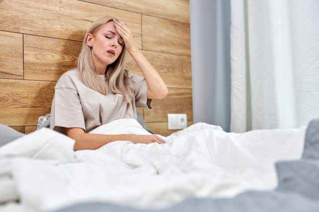Frau, die krank ist und Grippe hat, sitzt allein zu Hause im Bett, hat hohes Fieber oder Temperatur und berührt die Stirn