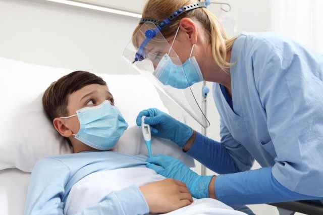 Krankenschwester mit Thermometer misst Fieber am Patientenkind im Krankenhausbett, trägt Schutzvisier und OP-Maske.
