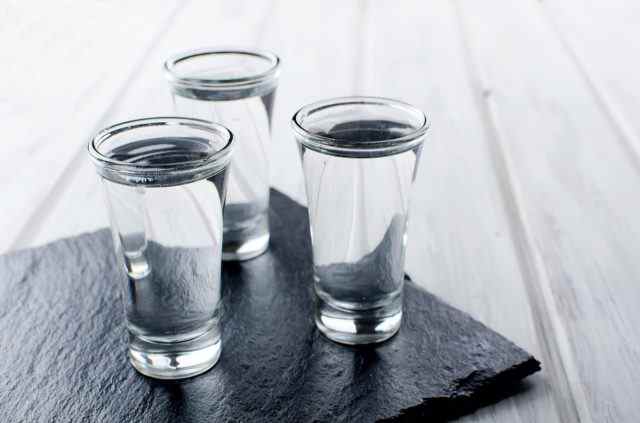 Drei Wodka-Shots mit Eis auf schwarzem Steinbrett auf weißem Holztisch, Kopierraum
