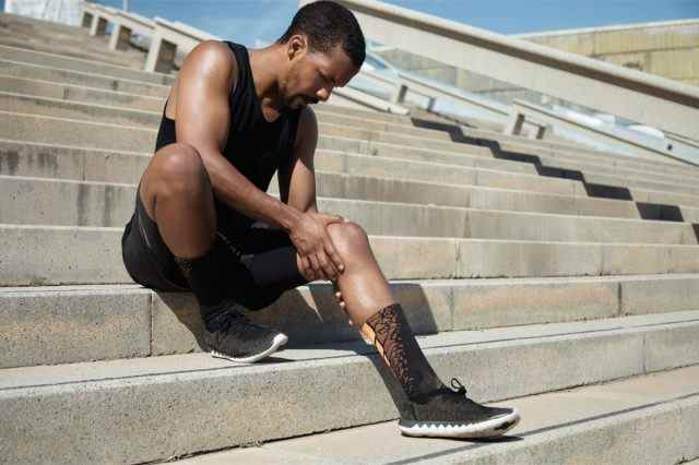 Schwarzer männlicher Jogger in schwarzer Sportbekleidung und Sportschuhen, der draußen auf der Treppe sitzt und sein schmerzendes Knie umklammert
