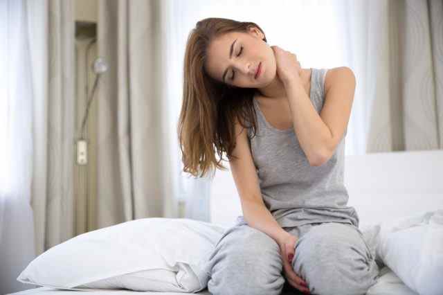 Frau sitzt mit Nackenschmerzen auf dem Bett