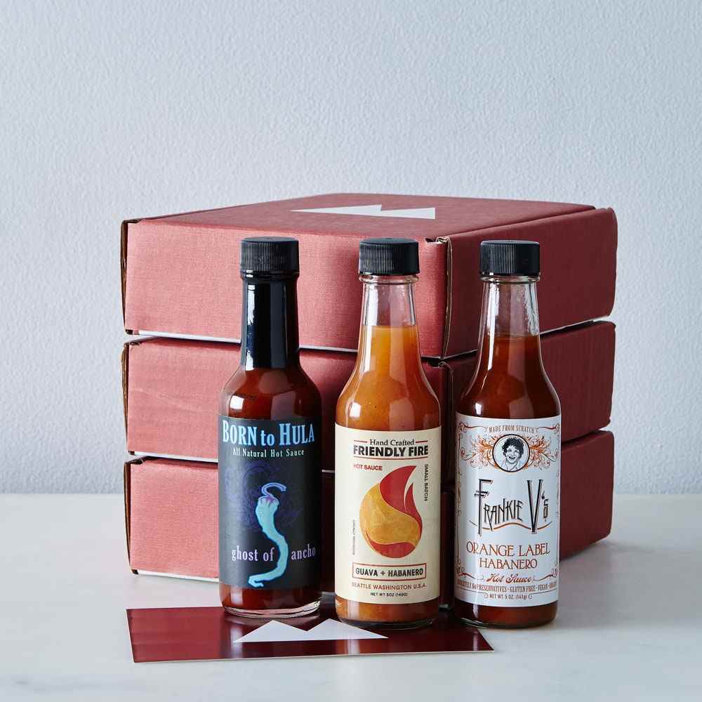 Drei verschiedene Hot-Sauce-Flaschen vor drei roten Food52-Boxen auf grauem Hintergrund