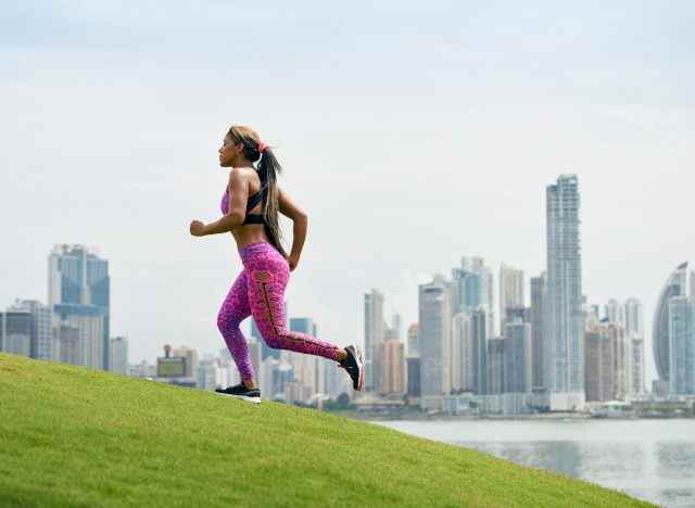Frau, die bergauf joggt, um doppelt so viele Kalorien zu verbrennen, sonniger Tag mit Stadtkulisse