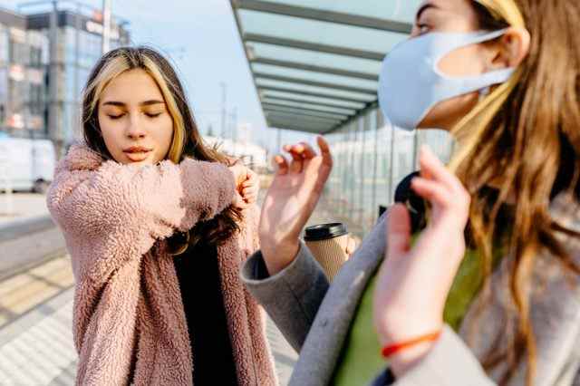 Junge kranke Studentin im Teenageralter draußen an der Bushaltestelle niest durch eine Allergie oder Erkältung in den Ellbogen.  Verängstigte Frau in Schutzmaske, verängstigte Hustenfrau im Freien
