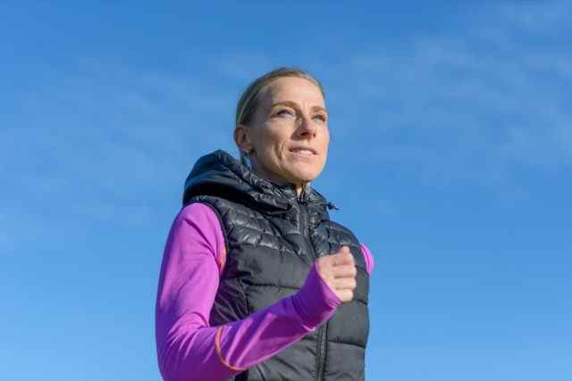 Frau mittleren Alters, die im Winter in einer Nahaufnahme mit niedrigem Winkel vor einem sonnigen blauen Himmel in einem gesunden, aktiven Lebensstil joggt