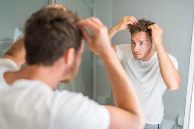 junger Mann in weißem T-Shirt und Jeans, der im Spiegel auf dünner werdendes Haar blickt