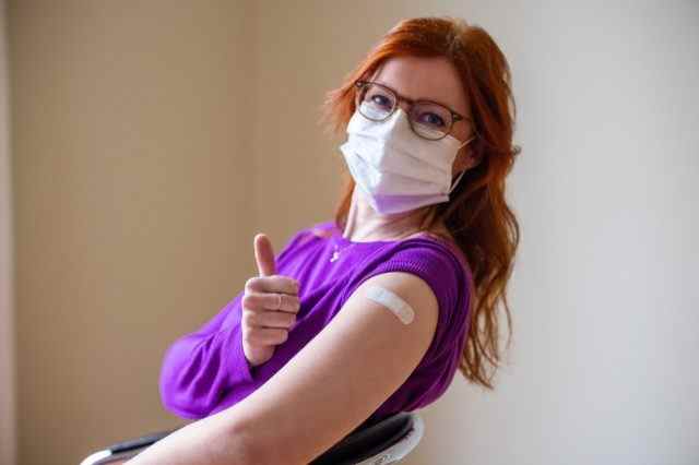 Frau mit Gesichtsmaske, die in die Kamera blickt und Daumen nach oben zeigt, nachdem sie den Covid-19-Impfstoff erhalten hat.