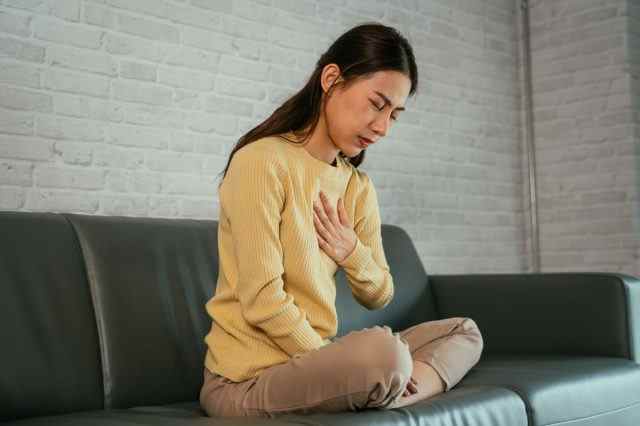 Asiatische junge Frau, die sich unwohl fühlt, weil sie an Sodbrennen leidet, die Brust mit geschlossenen Augen hält und mit gefalteten Beinen zu Hause auf der Couch sitzt.