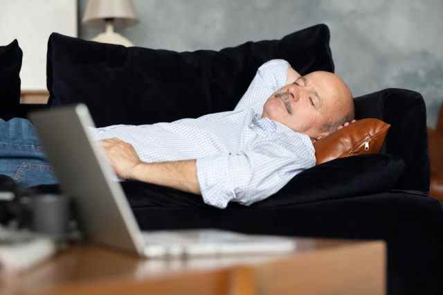 Müder älterer hispanischer Mann, der auf einer dunkelblauen Couch schläft und im Wohnzimmer ein Mittagsschläfchen hält