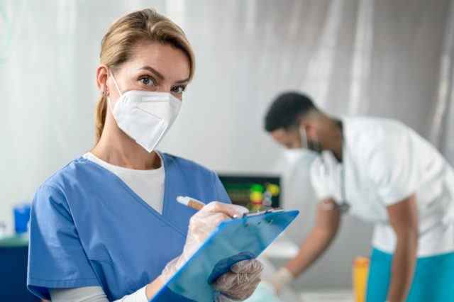 Porträt eines Arztes mit Gesichtsmaske und Klemmbrett, der im Krankenhaus in die Kamera schaut.